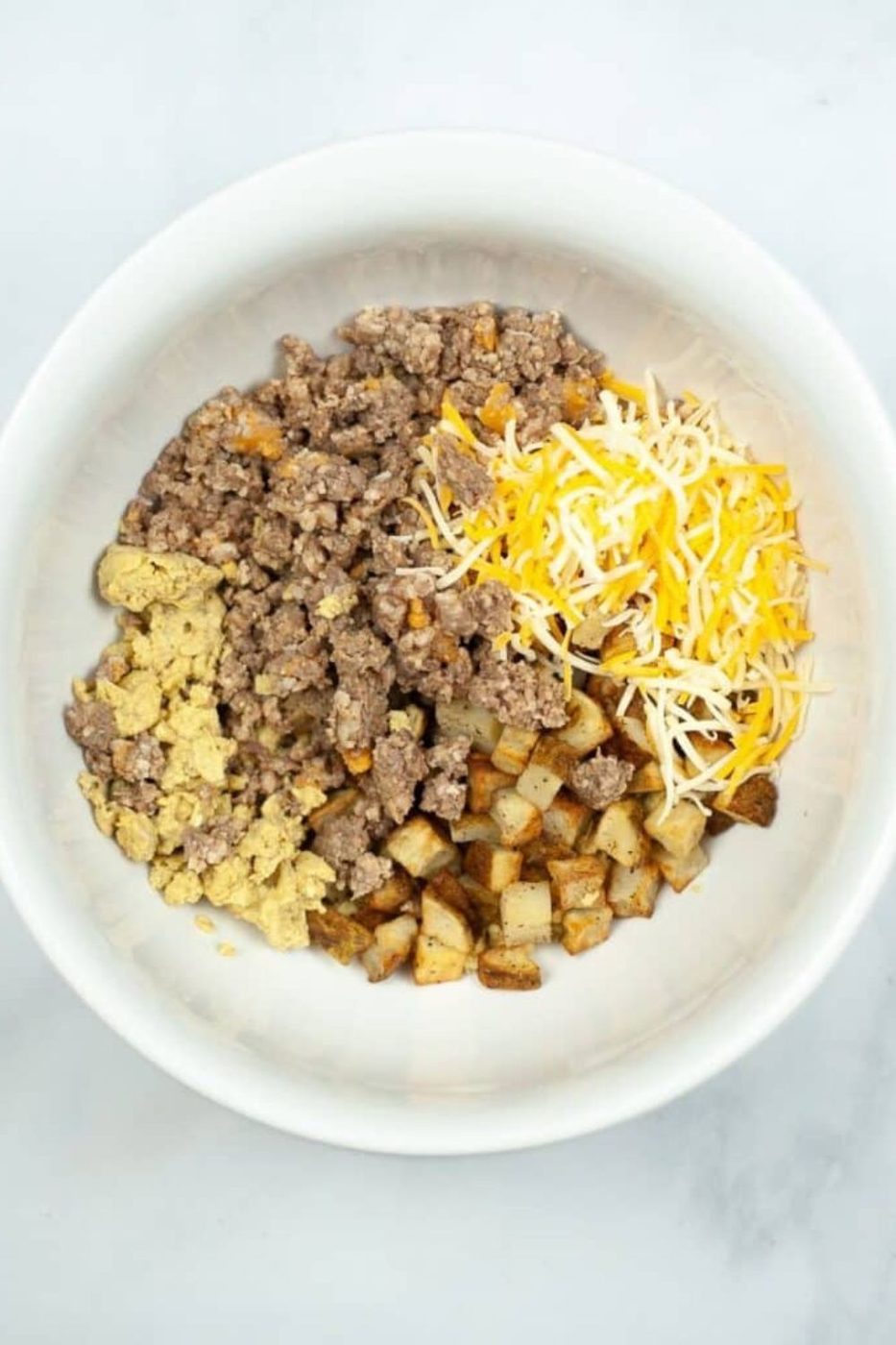 white bowl on table with cooked sausage, diced potatoes and shredded cheese in it
