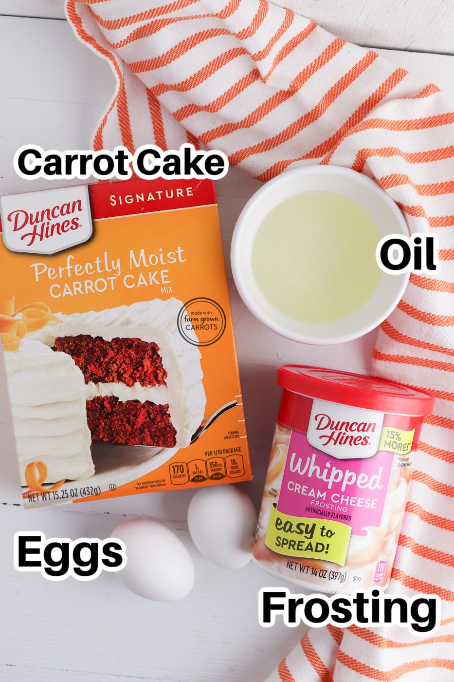 cake mix, oil in bowl, two eggs, and tub of frosting on white counter with a striped towel beside it