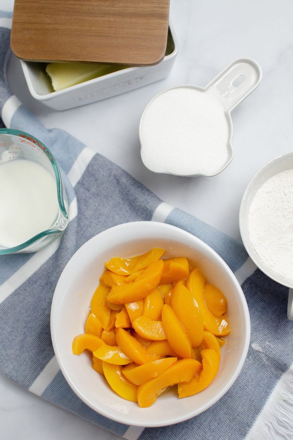 peaches in a bowl, flour, sugar and milk in bowls around the peaches on table 