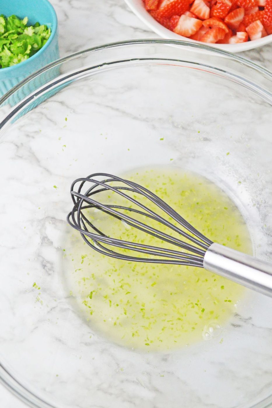 honey and lime juice mixed in a glass bowl with a whisk in bowl on marble counter top 