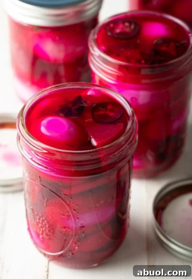 Three glass pint jars filled with vivid pink beet spicy pickled eggs, interspersed with chunks of beets, slices of fresh jalapeno, and whole star anise, creating an appealing and colorful display.