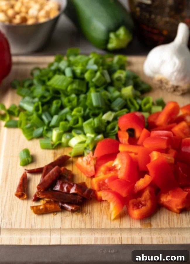 Freshly diced red bell peppers, vibrant green scallions, potent red chiles, and a bulb of garlic neatly arranged on a wooden cutting board, ready for prep.