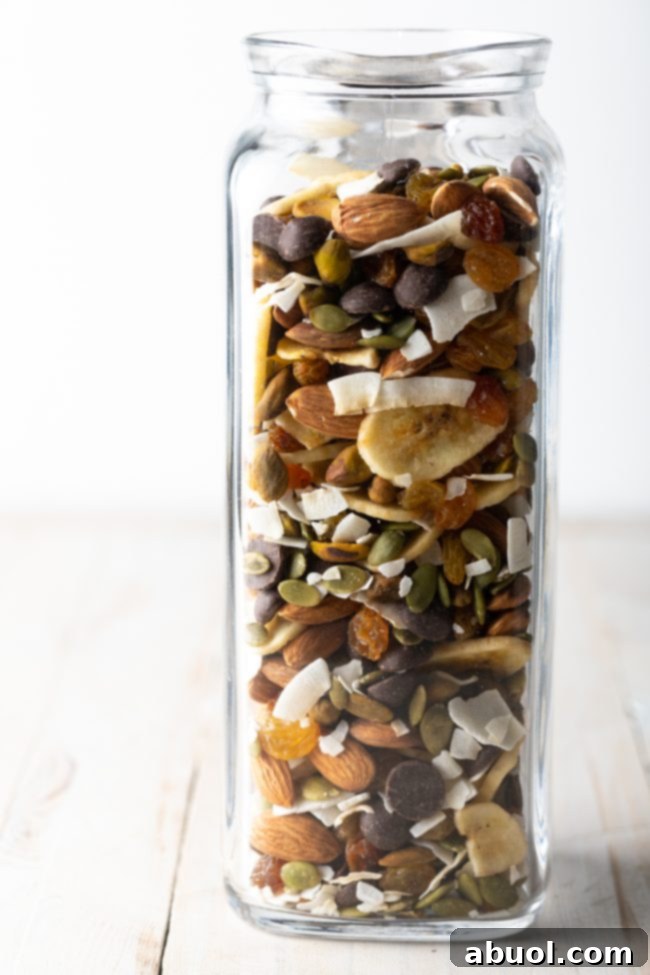 A side view of a glass jar filled with healthy homemade trail mix, showing layers of nuts, seeds, and dried fruit.