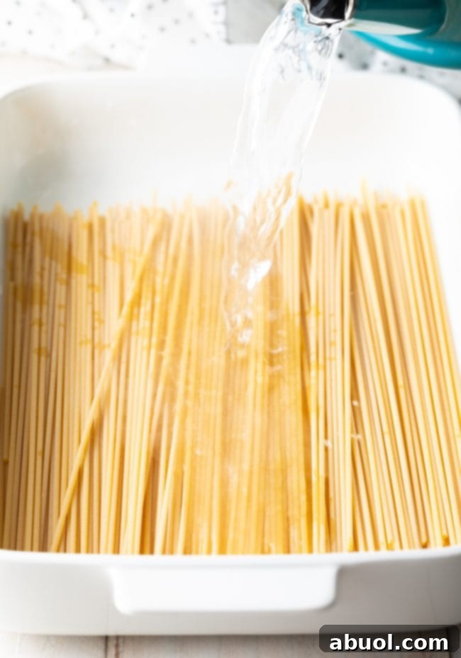 Pouring hot water over bucatini pasta in a white baking dish.