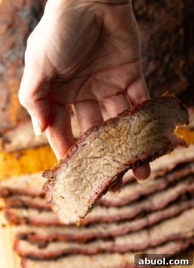 Dry-Brined Smoked Brisket 4 A hand holds up a succulent slice of smoked brisket, demonstrating its tenderness and a visible smoke ring. More sliced brisket is blurred in the background.