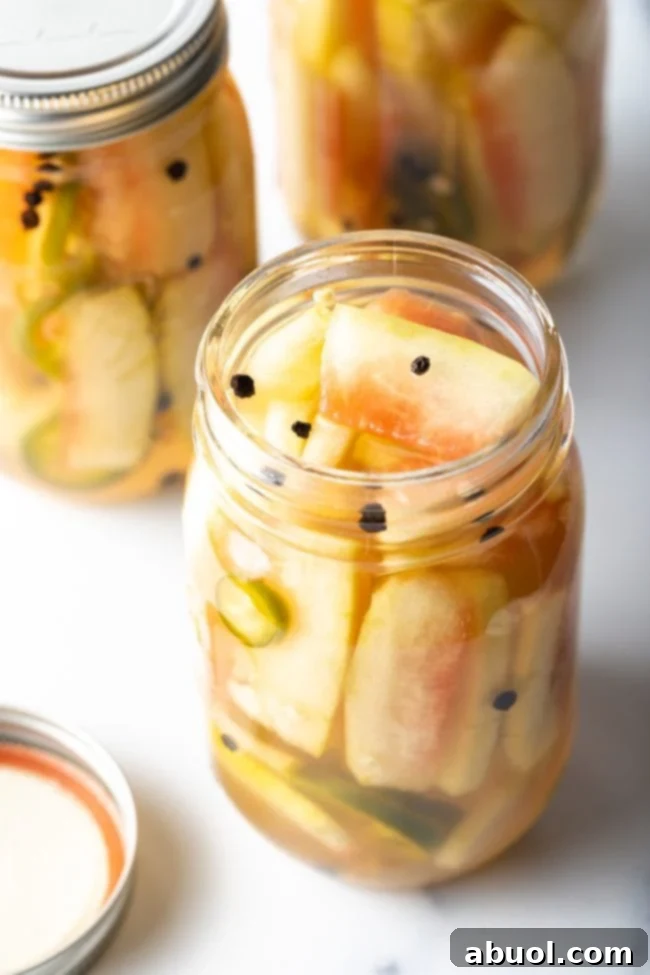 An open glass jar of pickled watermelon rinds, with visible peppercorns and jalapeño slices.