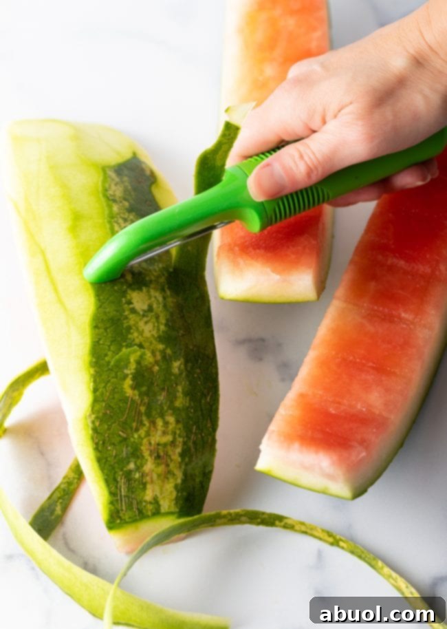 A hand expertly peeling the green outer layer off a segment of watermelon rind using a vegetable peeler.