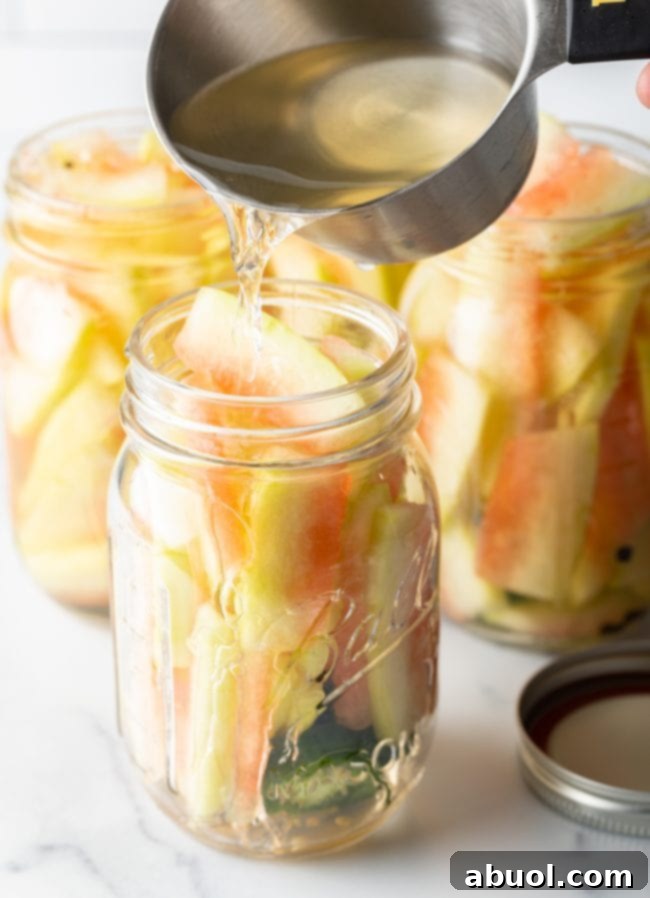 Hot pickling brine being carefully poured into jars filled with watermelon rinds, jalapeños, and peppercorns.