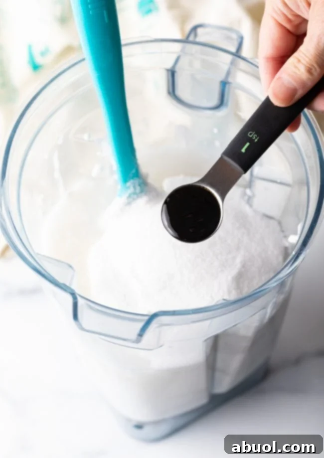 Vanilla extract being added to a blender filled with a creamy coconut milk mixture, ready for blending.