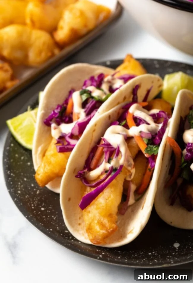 Three fish tacos on a black plate topped with slaw and served with lime wedges. 