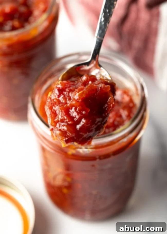 A jar of rich, red tomato jam with a spoon scooping out a generous portion, showcasing its thick, glossy texture.