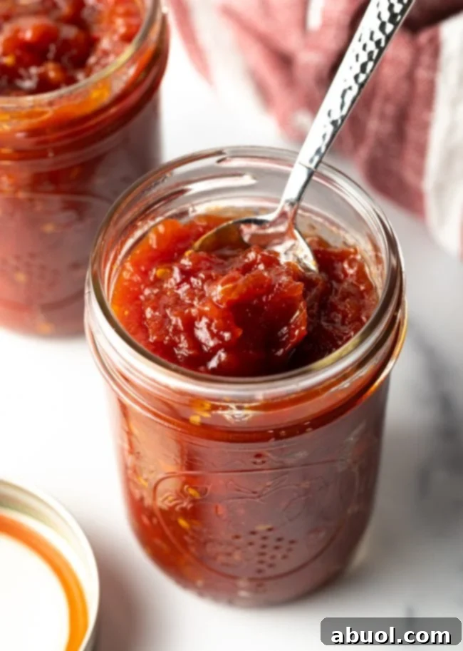 A hand spoons out a perfectly thick and glossy scoop of homemade tomato jam from a rustic mason jar.