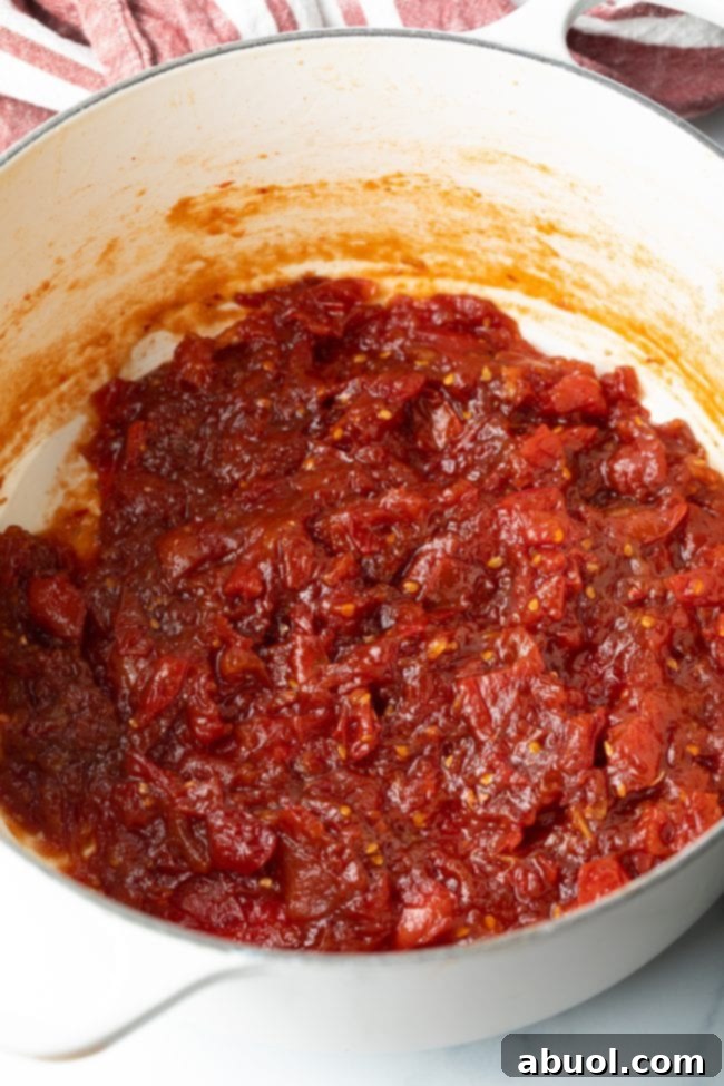 Tomato jam actively simmering in a pot on the stovetop, gradually thickening and developing rich color.