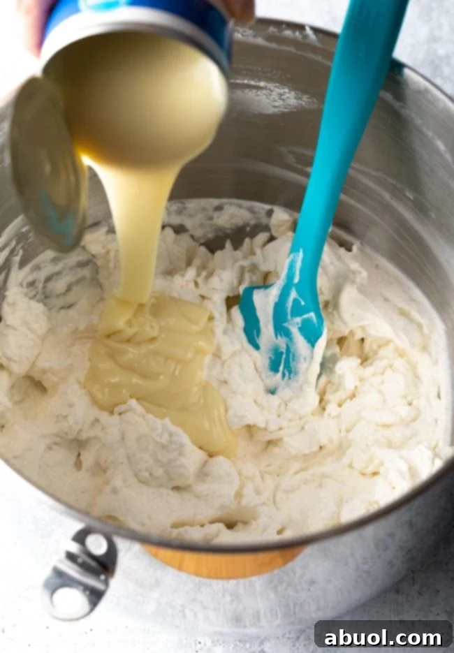 Rubber spatula gently folding sweetened condensed milk into fluffy whipped cream in a mixing bowl.