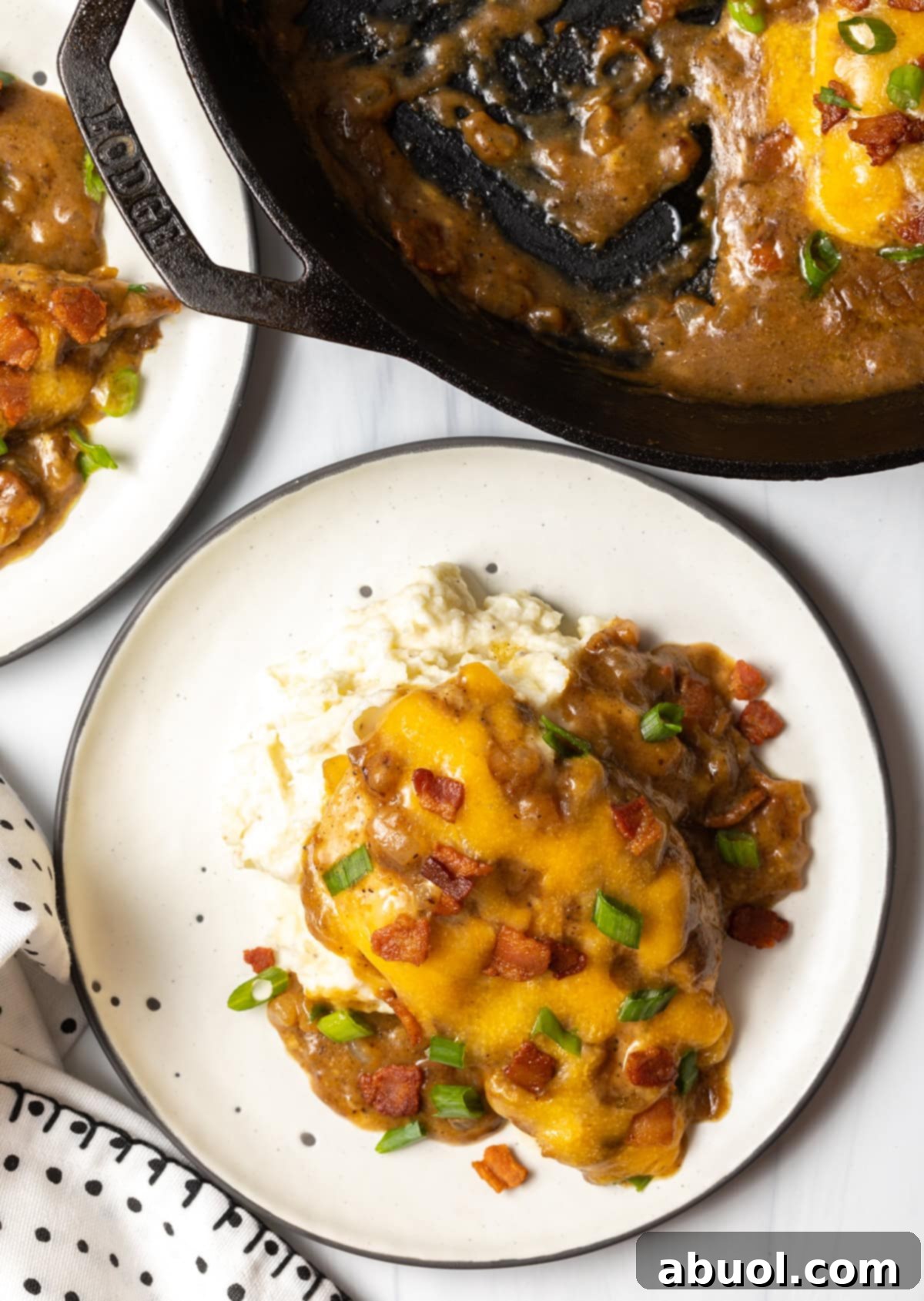 Overhead view of a white plate with a generous serving of mashed potatoes, accompanied by a chicken breast smothered in gravy and cheese, with the cast iron skillet holding more gravy visible in the background.