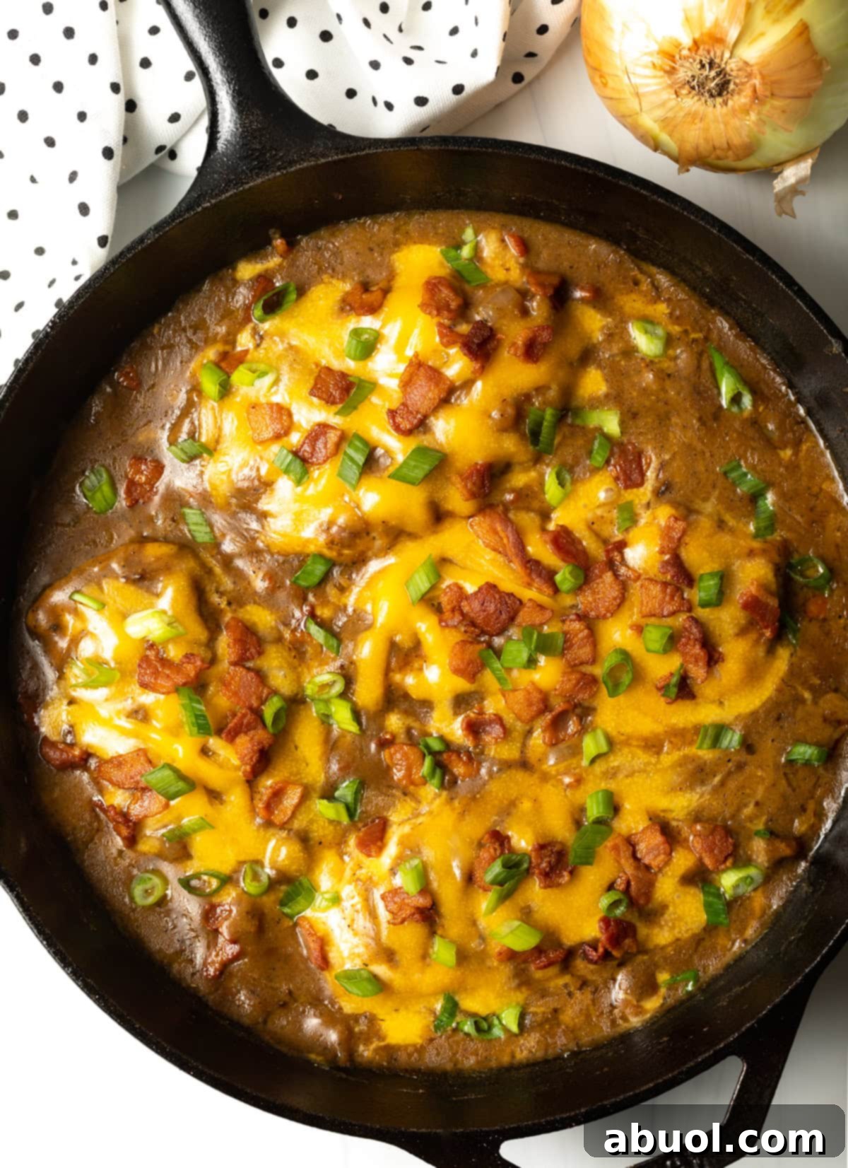 Overhead view of a cast iron skillet brimming with freshly cooked smothered chicken, golden-brown gravy, perfectly melted cheese, crispy bacon pieces, and bright green onions, ready to be served.