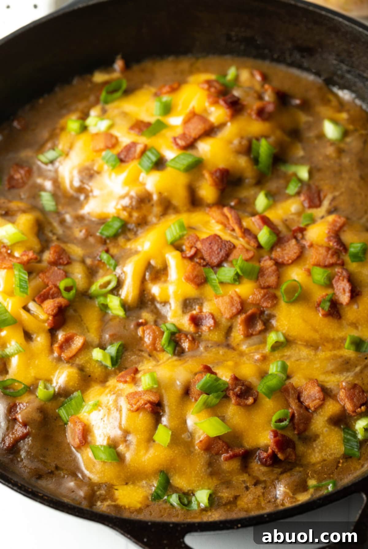 Close-up side view of a serving of smothered chicken and gravy recipe, topped with melted cheese, crispy bacon, and fresh scallions, presented beautifully in a cast iron skillet.