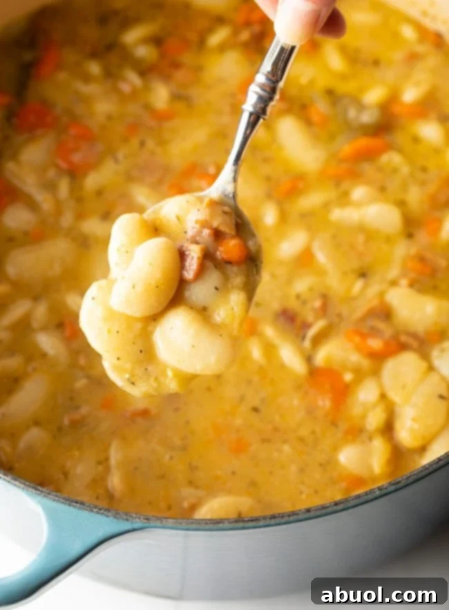 Spoon scooping lima beans, bacon, carrots, and aromatics from a large pot