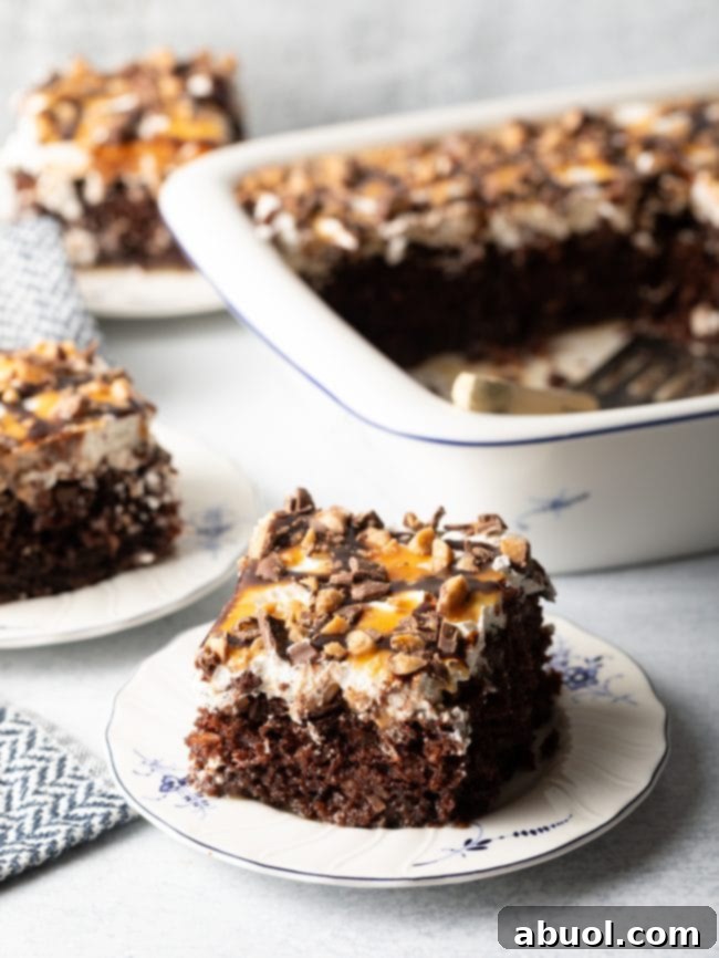 A slice of Better Than Sex Chocolate Poke Cake with whipped topping and caramel sauce on a white and blue plate, with the baking dish and spatula in the background