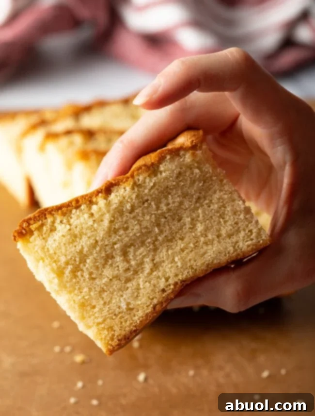 A hand gently holding a slice of moist Japanese wagashi yakimono, highlighting its soft texture, above a brown cutting board.