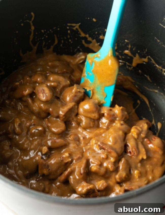 A blue spatula is actively stirring pecan halves into a rich, homemade caramel sauce within a large saucepot, ensuring every pecan is perfectly coated.