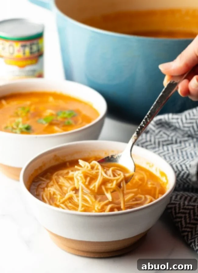 Hand holding a spoon, lifting a bite of Sopa de Fideo from a white and brown bowl, with a second bowl in the background.