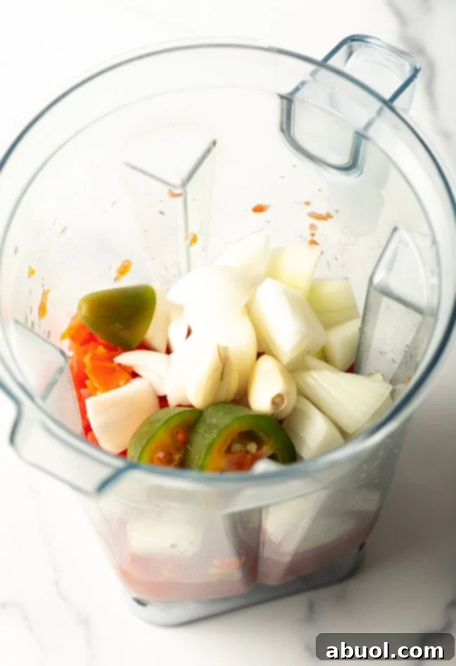 Top view of a blender jar with diced tomatoes, chopped onion, jalapeño, and garlic ready to be pureed.