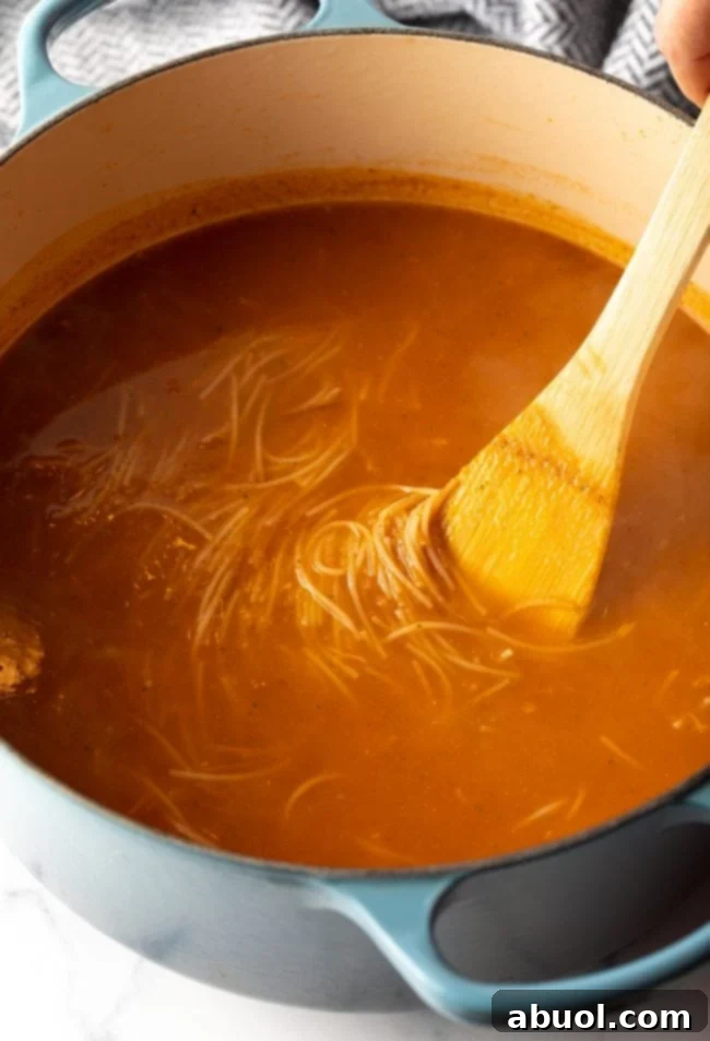 Wooden spoon stirring Mexican tomato soup with noodles in a large pot.