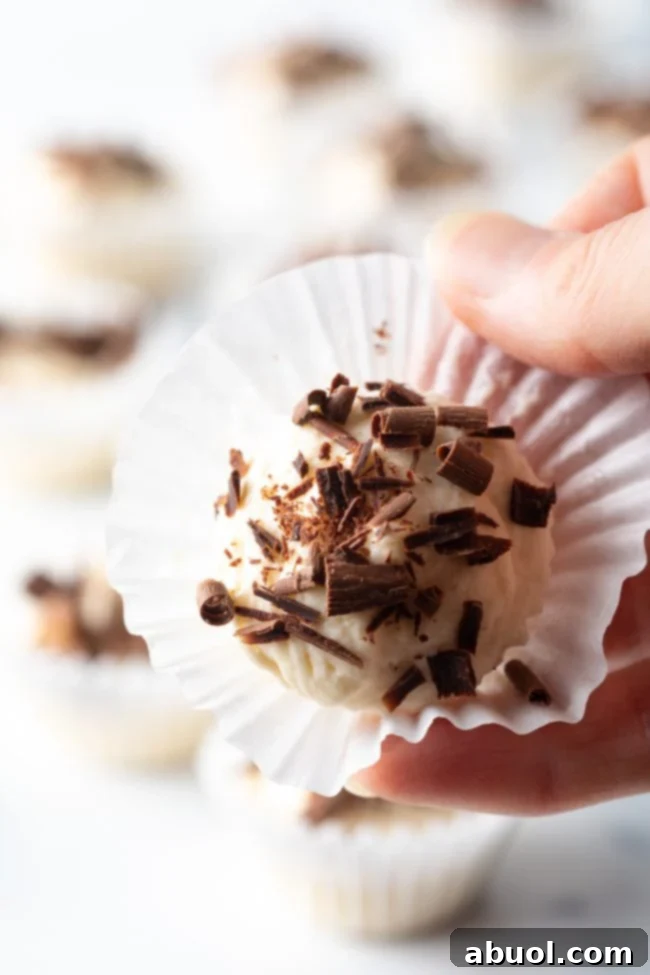 Hand holding full no-bake cheesecake fat bomb in a paper liner with chocolate shavings up to camera.
