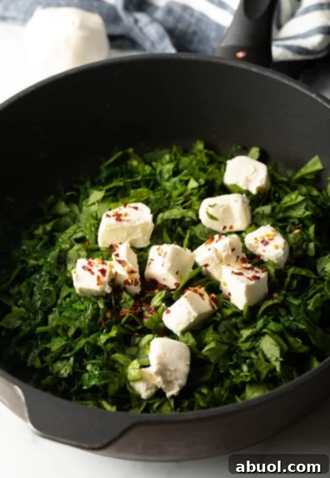 Cubed cream cheese and other sauce ingredients being added to the wilted spinach in a cast iron skillet.