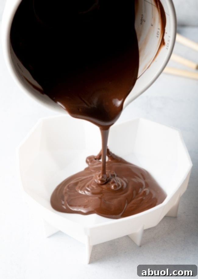 Melted chocolate being skillfully poured from a white measuring cup into a white silicone heart shell mold, demonstrating the precise coating process.