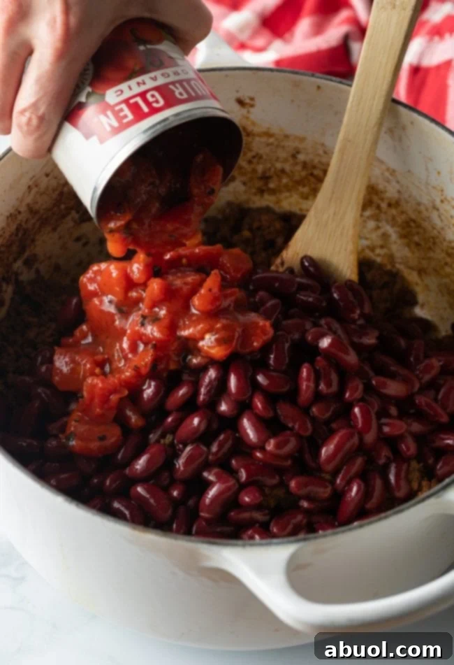 Hand adding canned tomatoes to kidney beans and beef in a large pot with wood spoon.