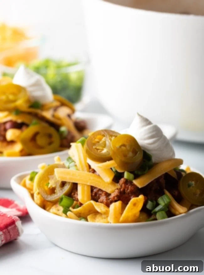Side view white shallow dish loaded with beef chili, corn chips, shredded cheese, chopped scallions, jalapeno slices, and a dollop of sour cream.