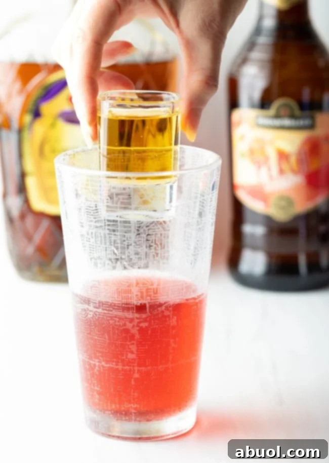 hand holding shot of red bull above the glass of whisky, cranberry juice, and peach schnapps