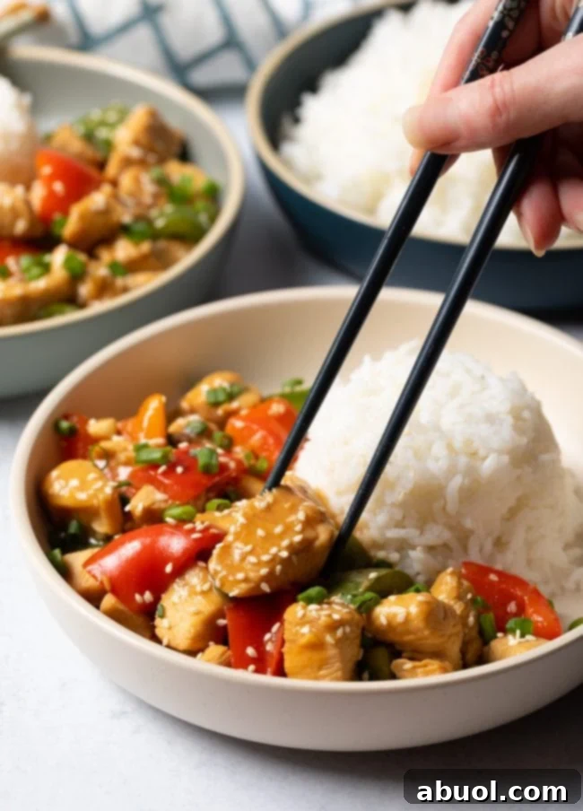 A hand holding chopsticks, picking up a tender piece of chicken from a bowl of Chinese garlic chicken stir-fry.