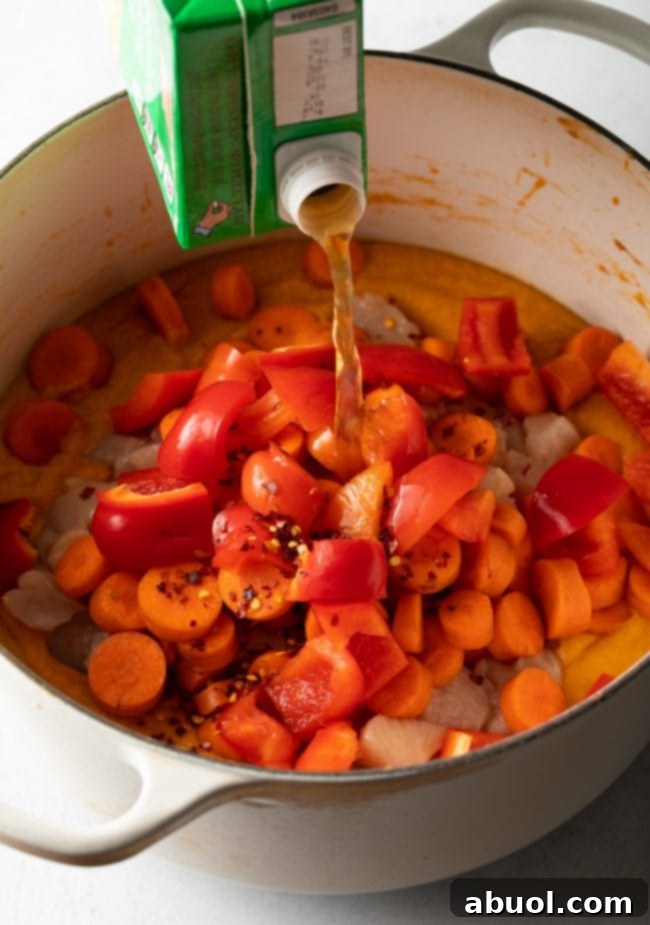 Chicken broth being poured into a pot over a simmering mixture of bell peppers, carrots, chicken pieces, and the creamy peanut butter base for Maafe.
