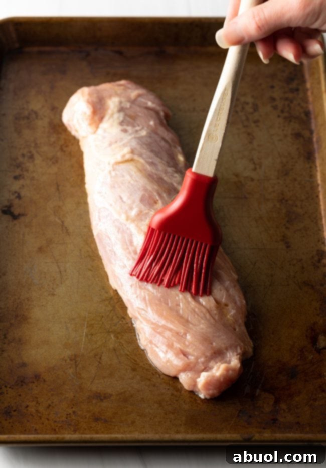 A basting brush applying oil to a raw pork tenderloin on a baking sheet, ready for seasoning.