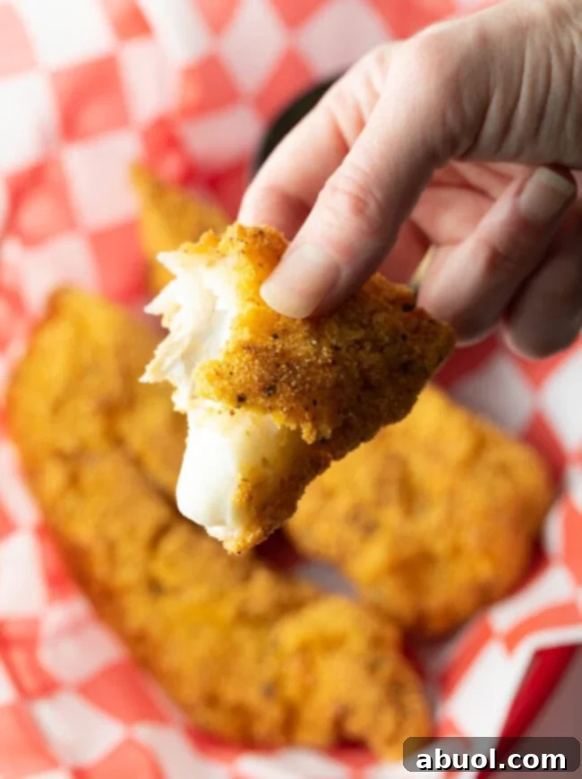A hand holding a piece of perfectly golden, crispy fried cornmeal fish towards the camera, showcasing its irresistible texture and inviting appeal.