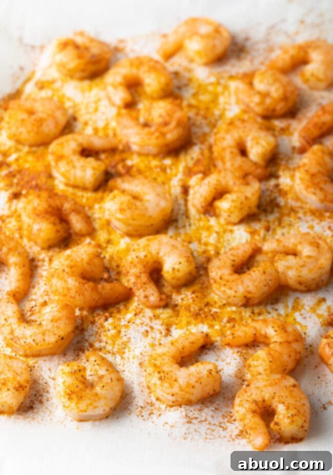 Oven-baked seasoned shrimp spread in a single layer on a parchment-lined baking sheet, showing their pink, curled 'C' shape.