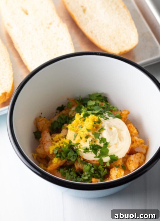 Cooked shrimp, mayonnaise, lemon zest, and chopped parsley combined in a glass mixing bowl, ready to be tossed.