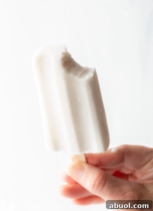 A hand holding a coconut ice pop with a bite taken out, showcasing its creamy texture.