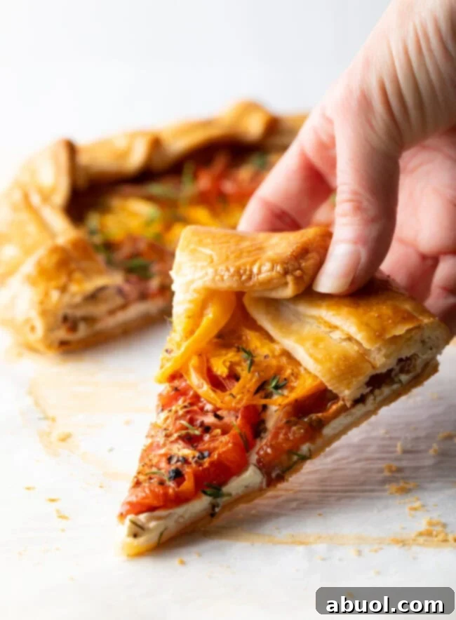 A hand holding a perfectly sliced piece of Italian Tomato Pie, showcasing its golden flaky crust and colorful layers of heirloom tomatoes and goat cheese.