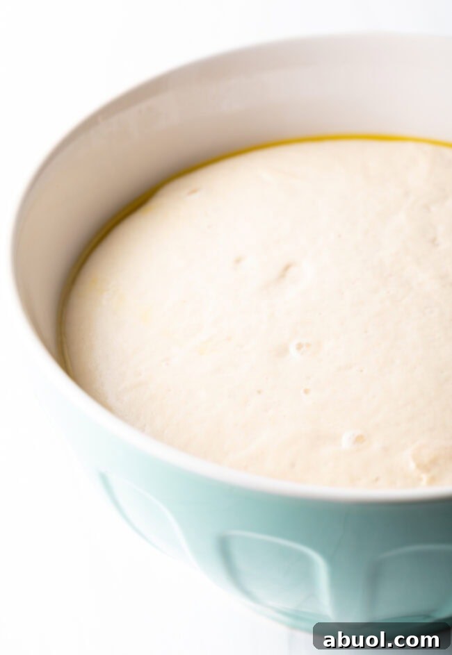 Close-up of focaccia dough actively proofing and rising in a large glass bowl, covered with plastic wrap.