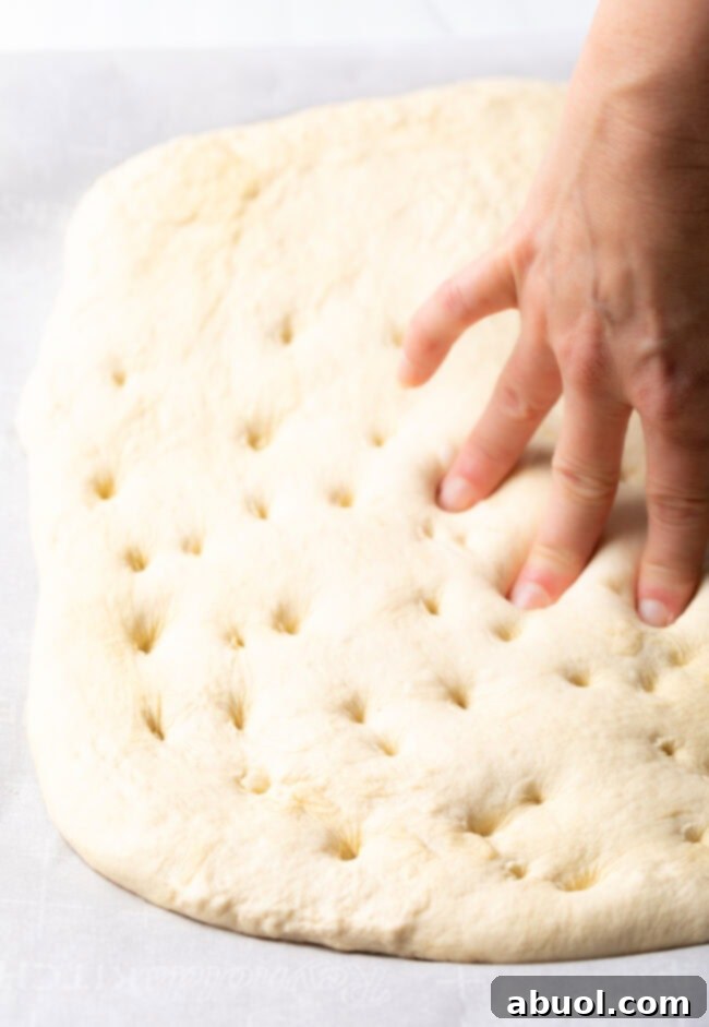 A hand gently pressing fresh basil leaves into the dimpled focaccia dough.