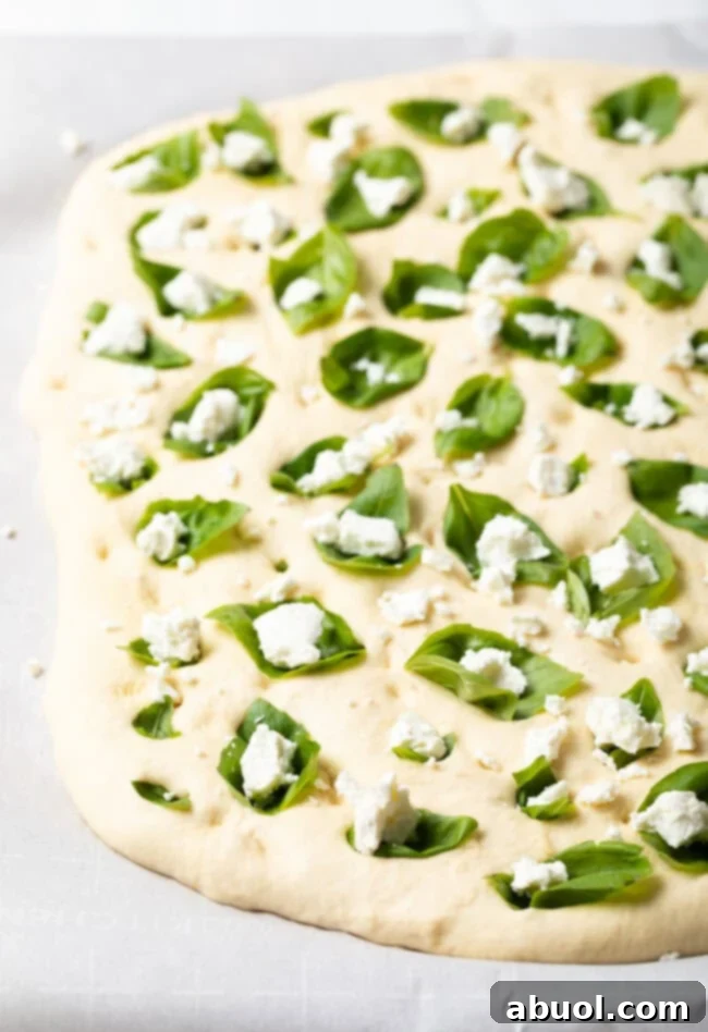 A hand pressing juicy cherry tomatoes into the dimples of the focaccia dough alongside basil and goat cheese.