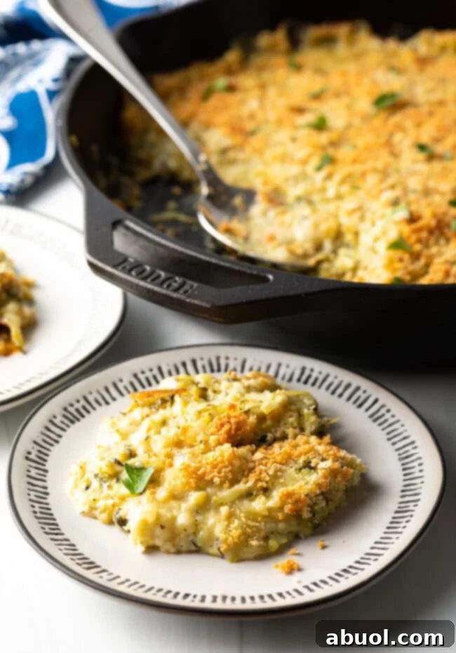 A delicious serving of cheesy zucchini casserole on a plate, with a cast iron skillet full of casserole in the background.