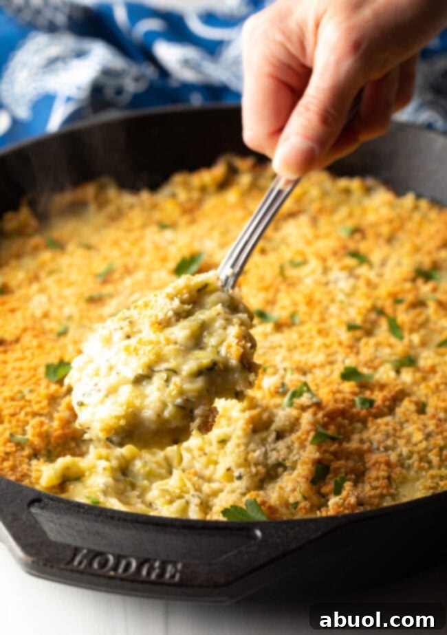 A hand using a metal spoon to scoop out a serving of zucchini casserole from a cast iron skillet.