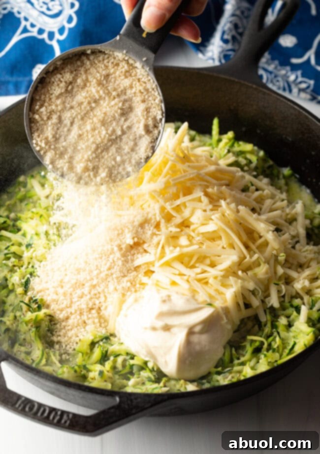 A hand adding Panko breadcrumbs from a measuring cup to the skillet containing zucchini, mayonnaise, and shredded cheese.