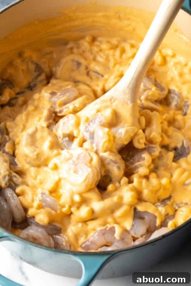 Wood spoon stirring cheesy mac and cheese and uncooked shrimp in a cooking pot.