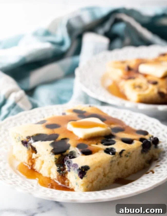 Fluffy blueberry sheet pan pancake square on a white plate, drizzled with butter and maple syrup. A perfect oven baked pancake breakfast.
